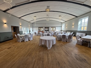 A large hall with round tables and chairs, high ceilings with chandeliers, and large windows.