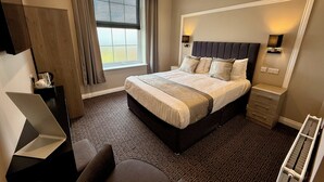 A hotel room with a large bed, a desk, a chair, a window with curtains, and a radiator.