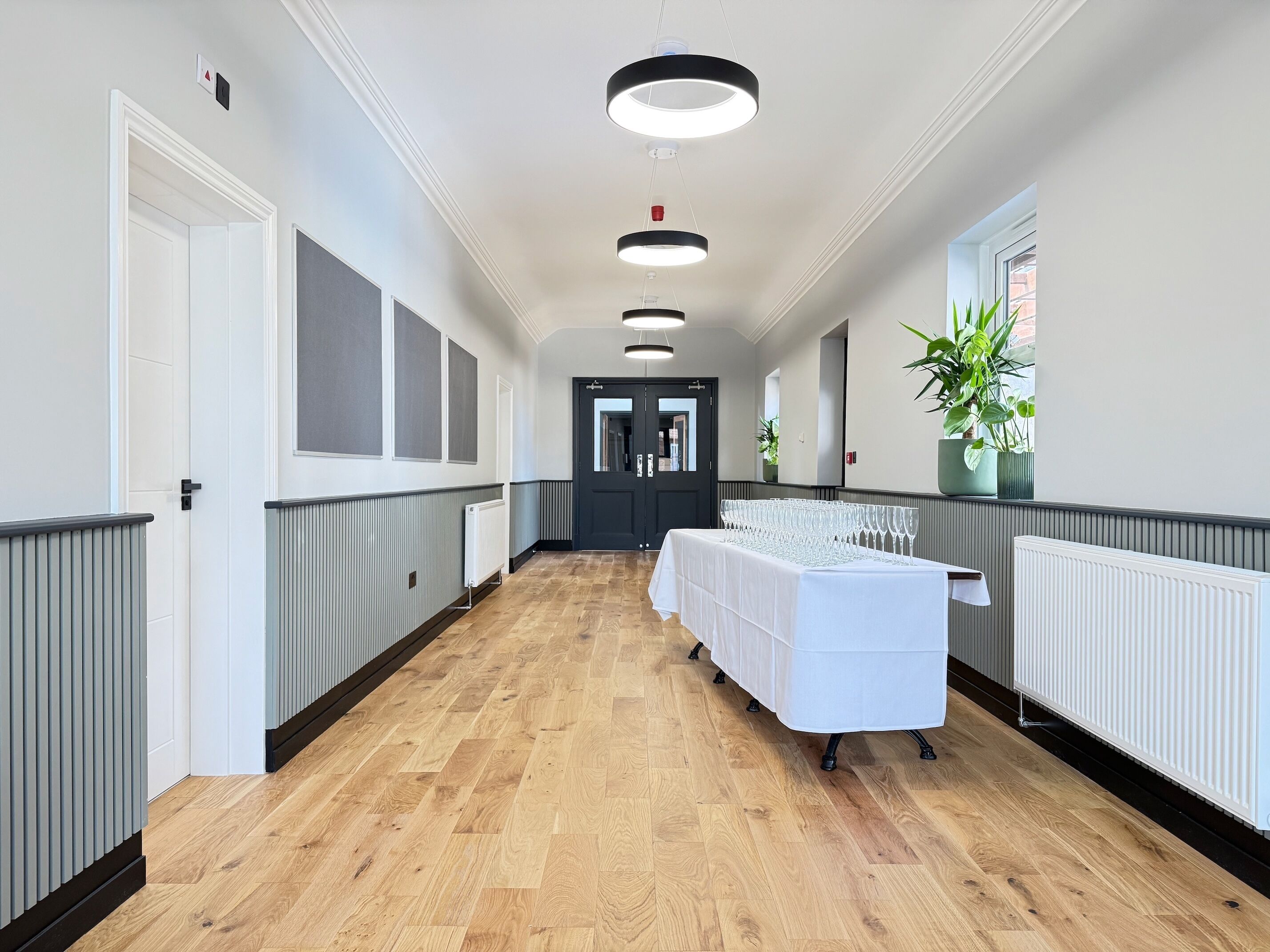 A hallway with a long table set with wine glasses, flanked by radiator panels and a potted plant.