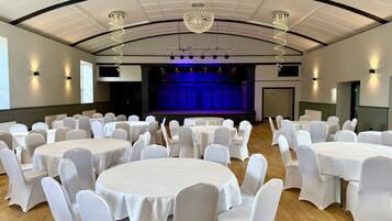 A banquet hall with round tables and white chairs, a stage with blue lighting, and chandeliers.