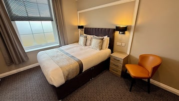 Quarto de hotel com uma cama grande, uma mesa de cabeceira, uma cadeira e uma janela com persianas.