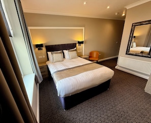 Quarto de hotel com uma cama grande, uma cadeira, um espelho e um radiador.
