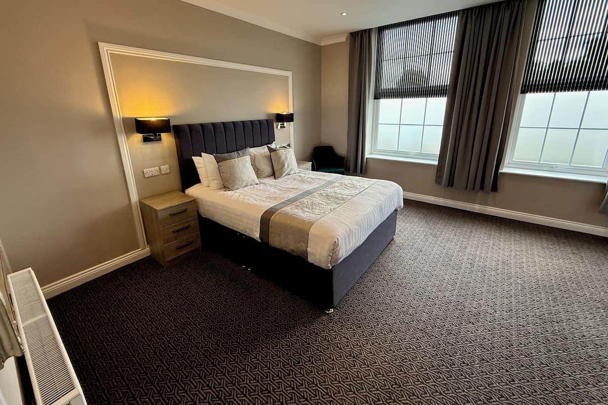 A hotel room with a large bed, bedside lamps, a nightstand, a window with curtains, and a patterned carpet.