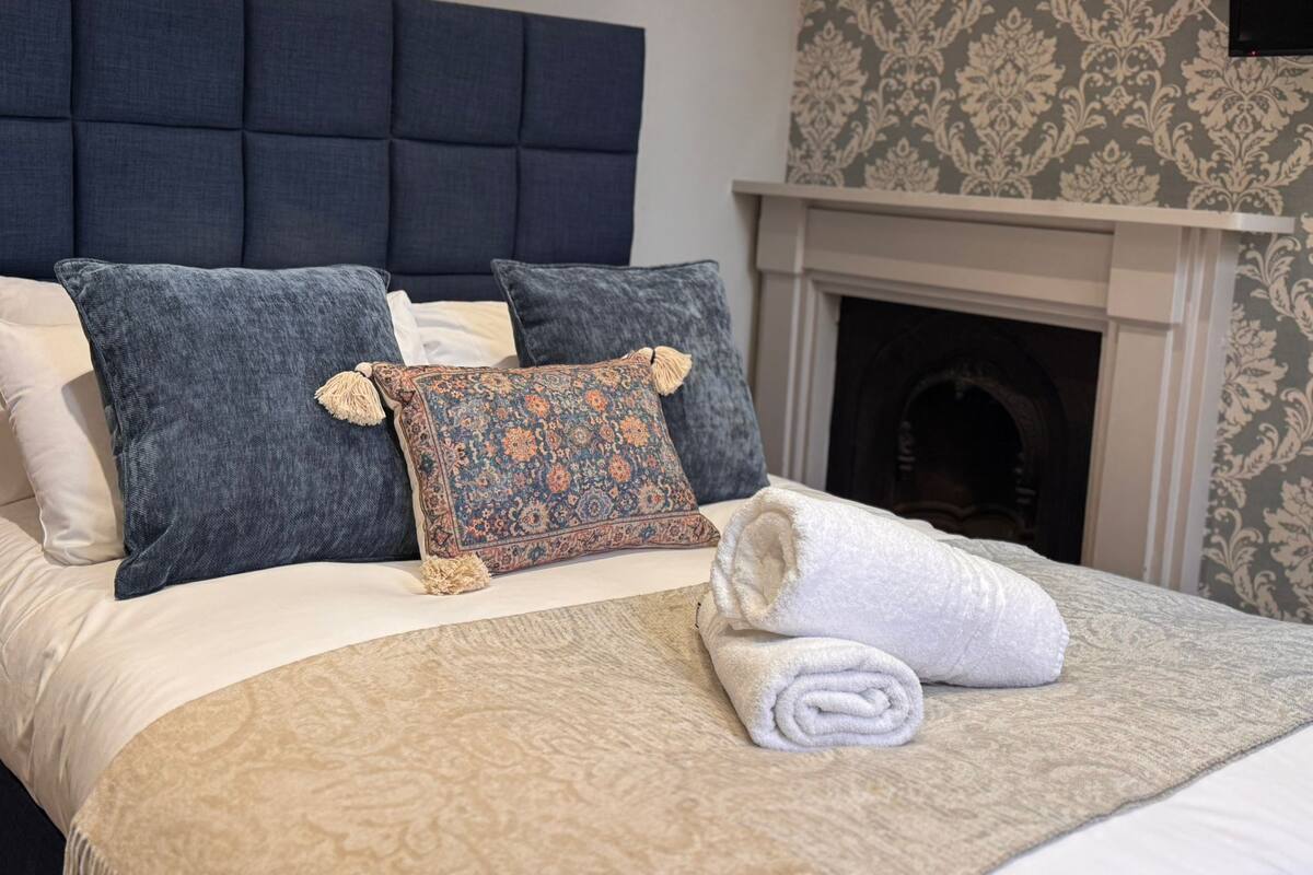 Una cama bien hecha con un plaid estampado, dos almohadas y una toalla enrollada.