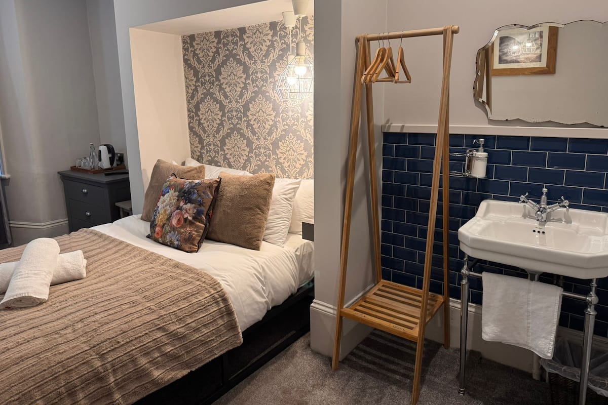 Un dormitorio con cama, un estante de madera para colgadores, un baño con lavabo y espejo, y un papel tapiz estampado.