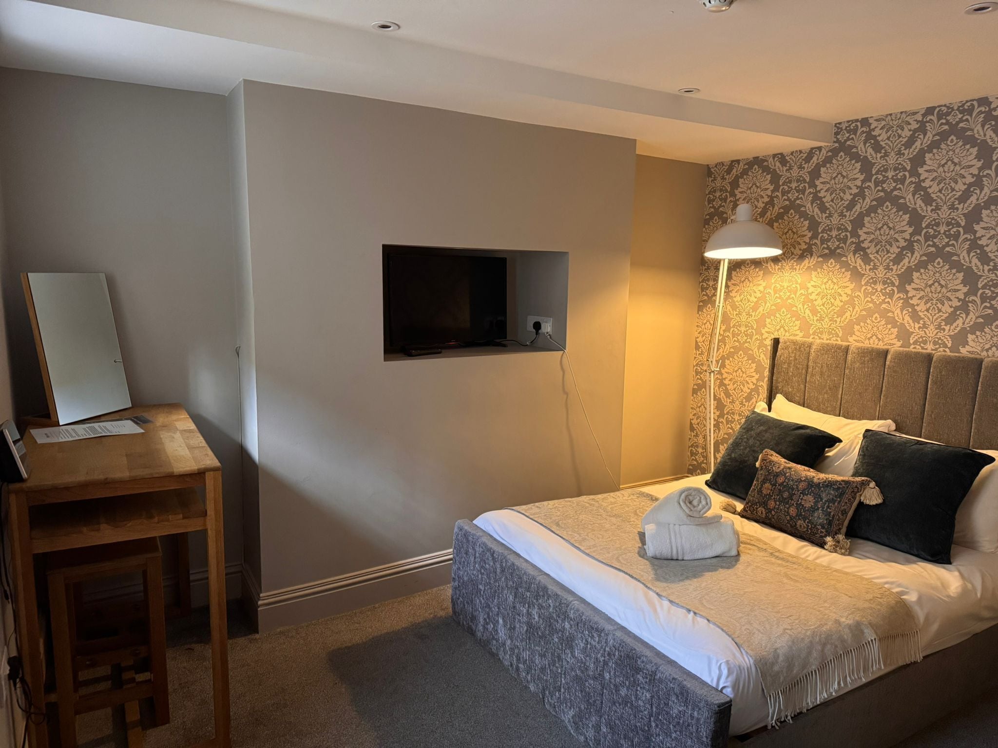A bedroom with a bed, a desk, a TV mounted on the wall, and a lamp.
