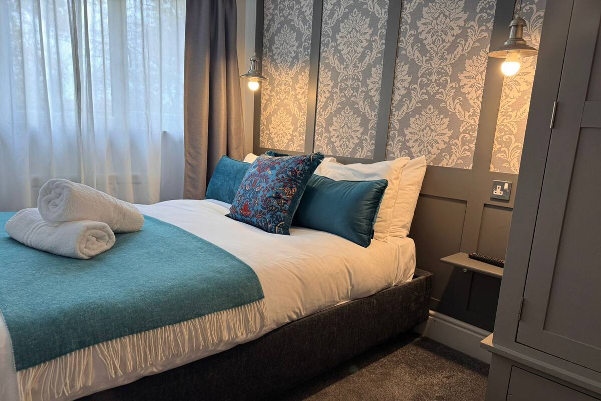 Una cama bien tendida con un edredón verde azulado, dos toallas enrolladas y almohadas estampadas.