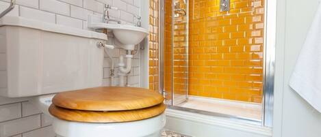 A bathroom with a white toilet, a wooden seat, a white sink, and a shower with a brick wall.