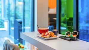A breakfast setup with a red bowl, a plate with food, a mug, and a pair of glasses on a white table by a window with a view of greenery. - ibis Styles Paris La Defense Courbevoie (Courbevoie)