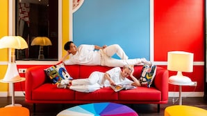 A red sofa with two people lying on it, a colorful wall, and two lamps. - ibis Styles Paris La Defense Courbevoie (Courbevoie)