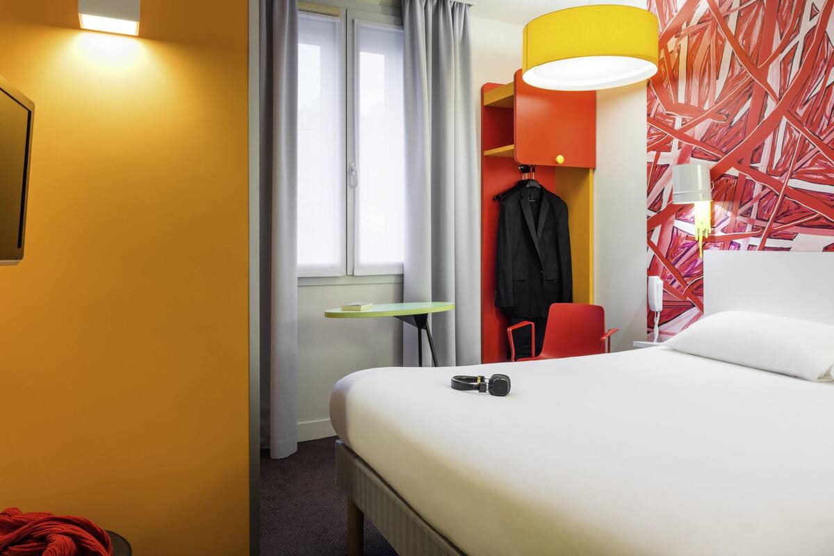 Een hotelkamer met een bed, een bureau, een stoel, een kapstok en een schilderij aan de muur.