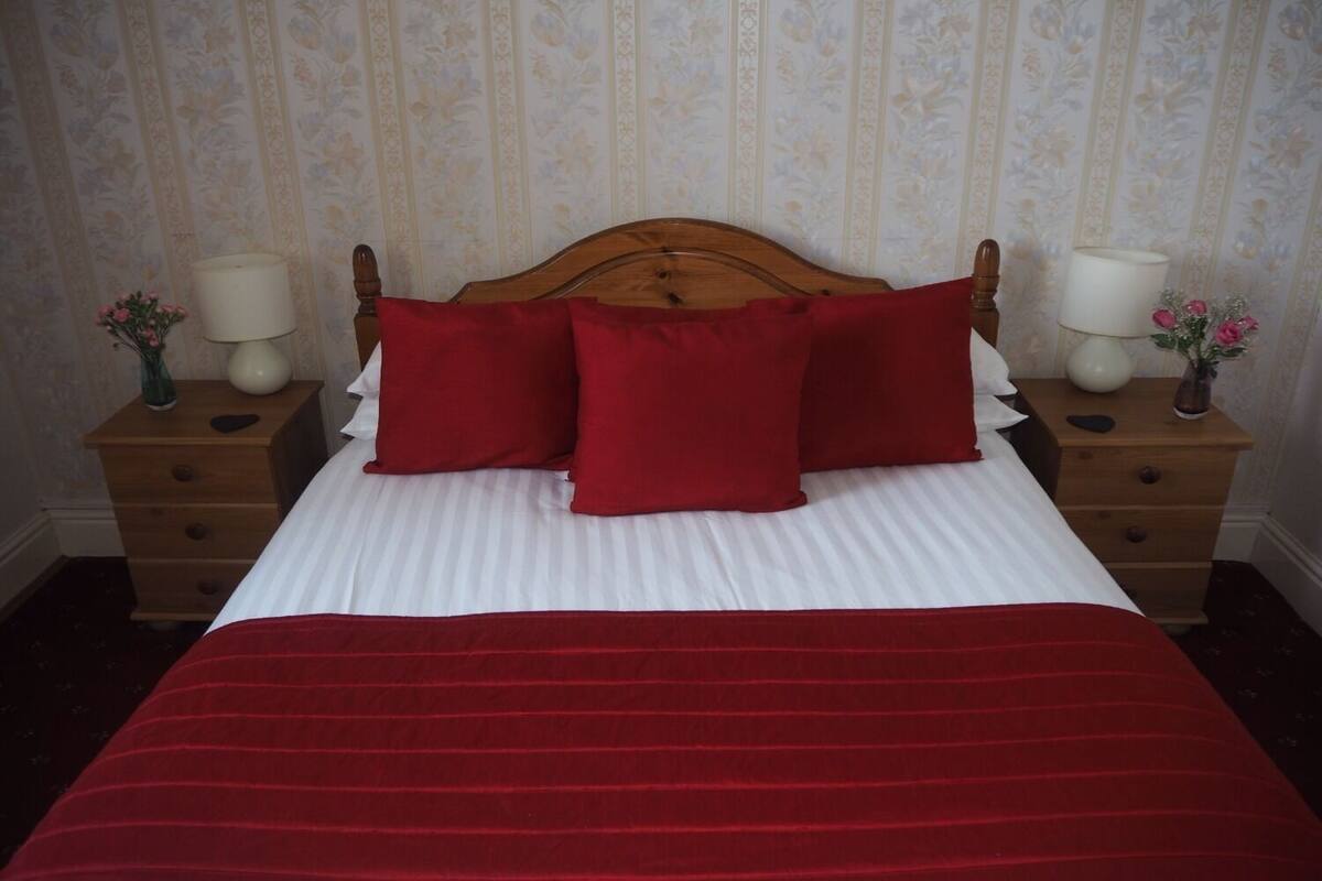 A neatly made bed with a red bedspread and pillows, flanked by wooden nightstands with lamps and vases of flowers.
