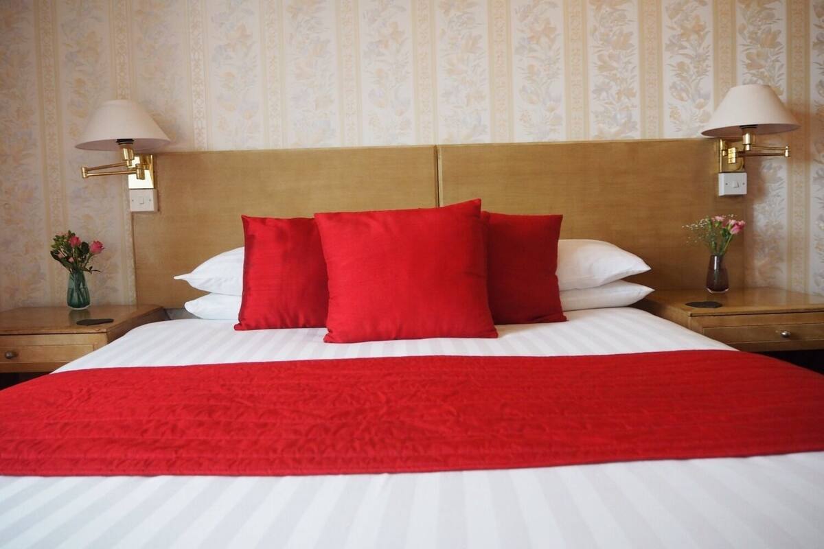 A hotel room with a large bed, red and white bedding, two nightstands with lamps, and floral wallpaper.