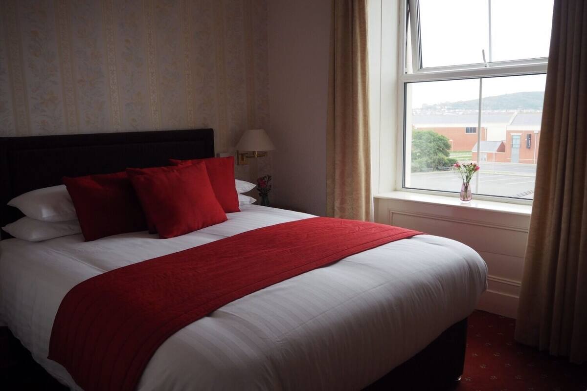 A hotel room with a large bed, red and white bedding, a window with a view of buildings, and a vase with flowers on the windowsill.