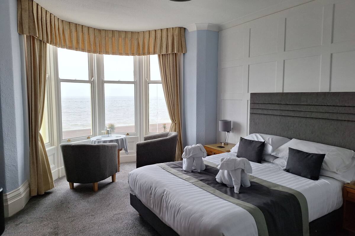 A hotel room with a large bed, two armchairs, a small table, and a view of the sea through large windows.