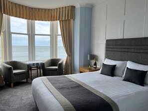 A hotel room with a large bed, two armchairs, a small table, and a view of the sea through large windows. - Swn Y Mor Hotel (Llandudno)