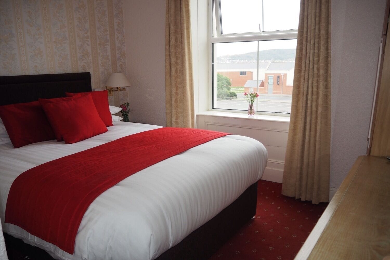 A hotel room with a large bed, red bedding, a window with curtains, and a view of buildings outside.