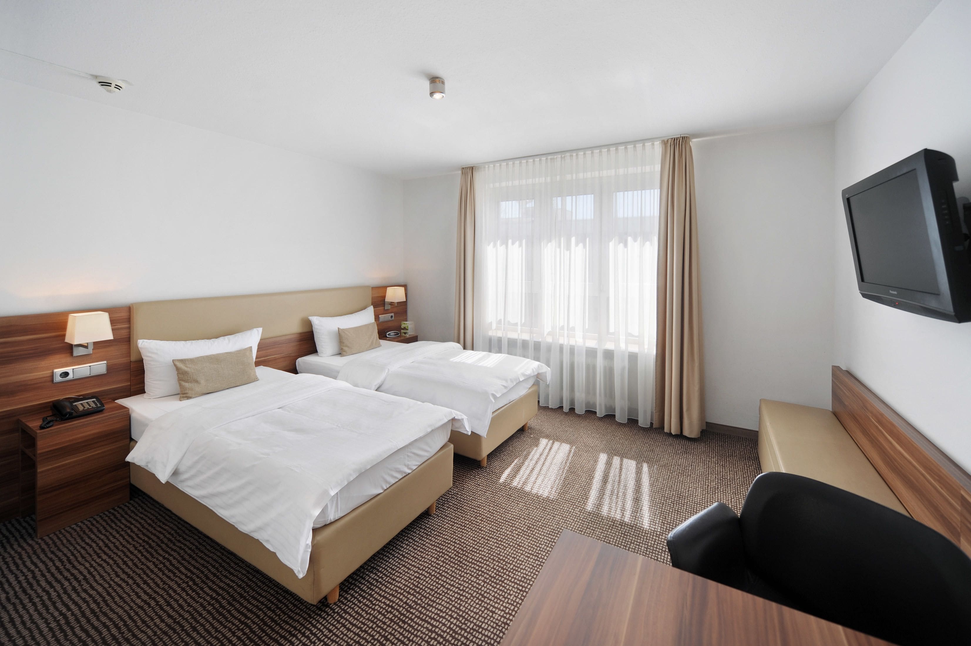 A hotel room with two beds, a desk, a chair, a TV, and a window with curtains.