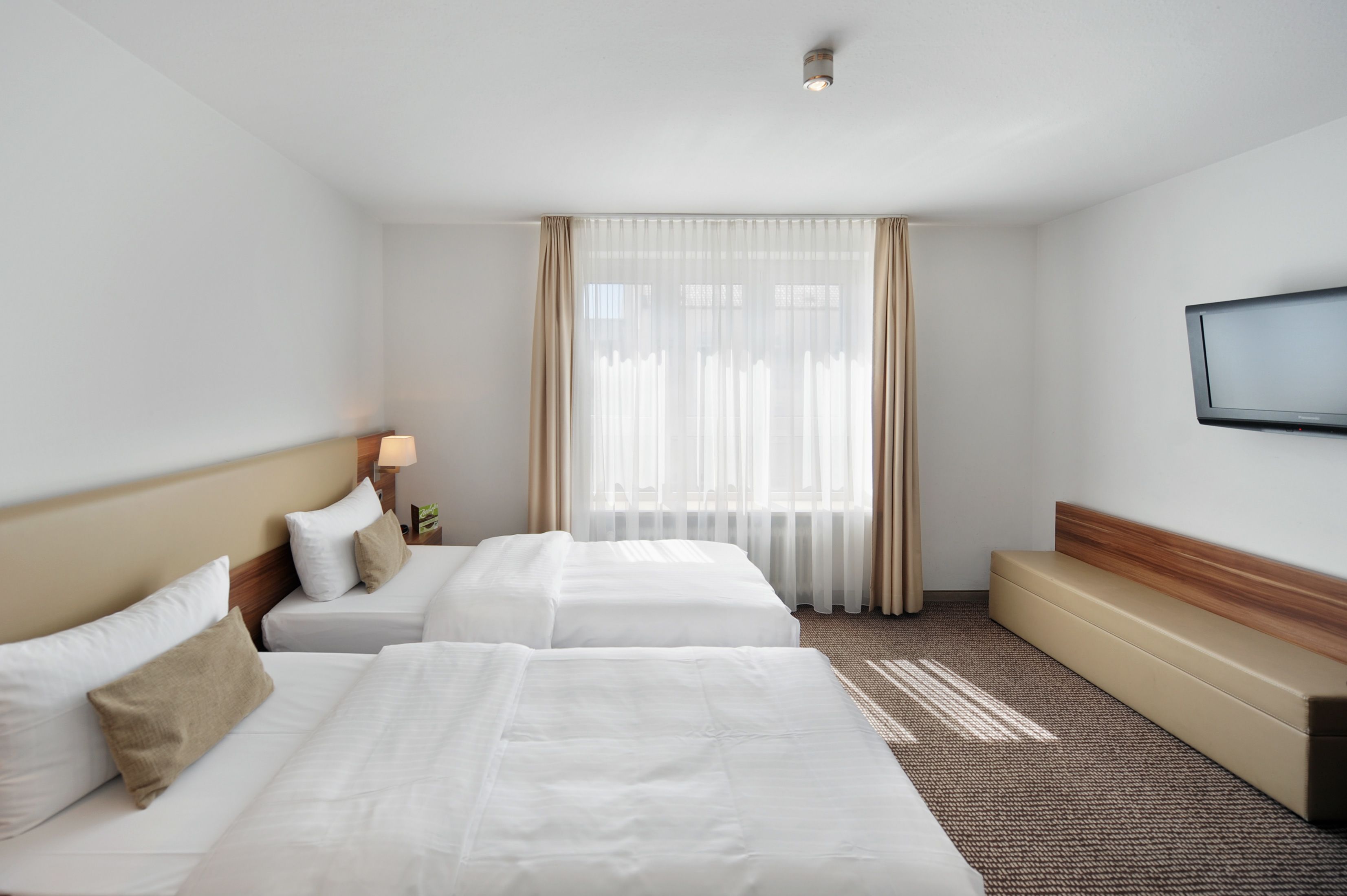 A hotel room with two beds, a flat-screen TV mounted on the wall, and a large window with sheer curtains.