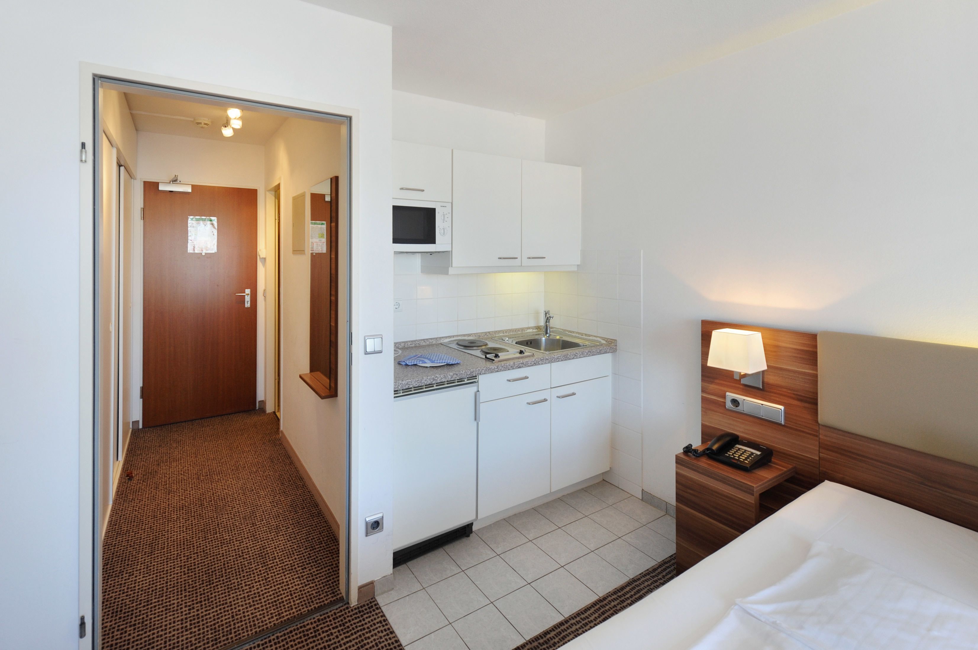 Een compacte hotelkamer met een kitchenette, een bed en een nachtkastje met lamp.
