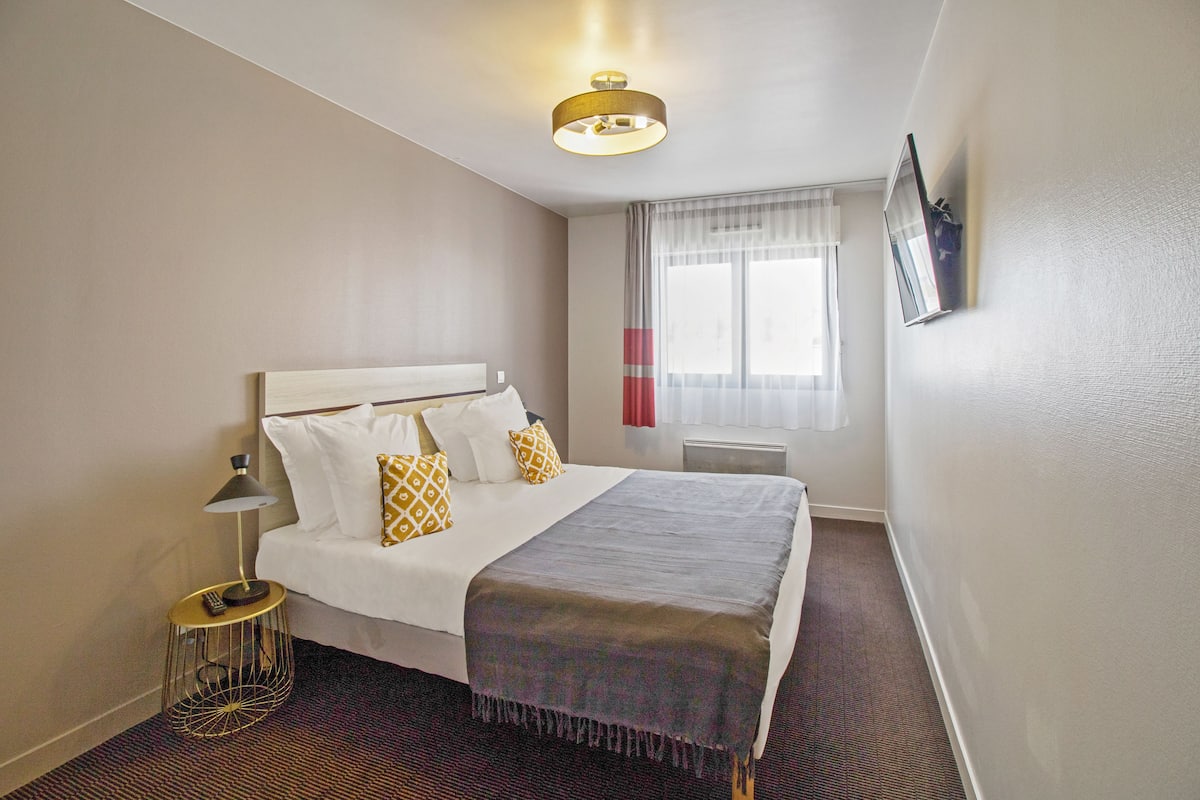 Habitación de hotel con cama, mesita de noche con lámpara, ventana con cortinas y televisor montado en la pared.