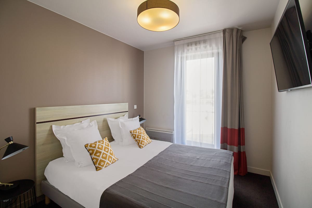 Habitación de hotel con una cama grande, una mesita de noche, una lámpara, un televisor y una ventana con cortinas.
