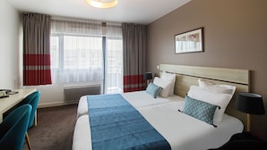 Habitación de hotel con cama, escritorio y silla. Tiene una ventana con cortinas, un cuadro colgado en la pared y una lámpara en el techo.