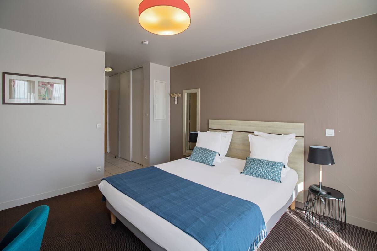Una habitación de hotel con una cama grande, una mesita de noche con lámpara, un cuadro enmarcado en la pared y una puerta que da a otra estancia.