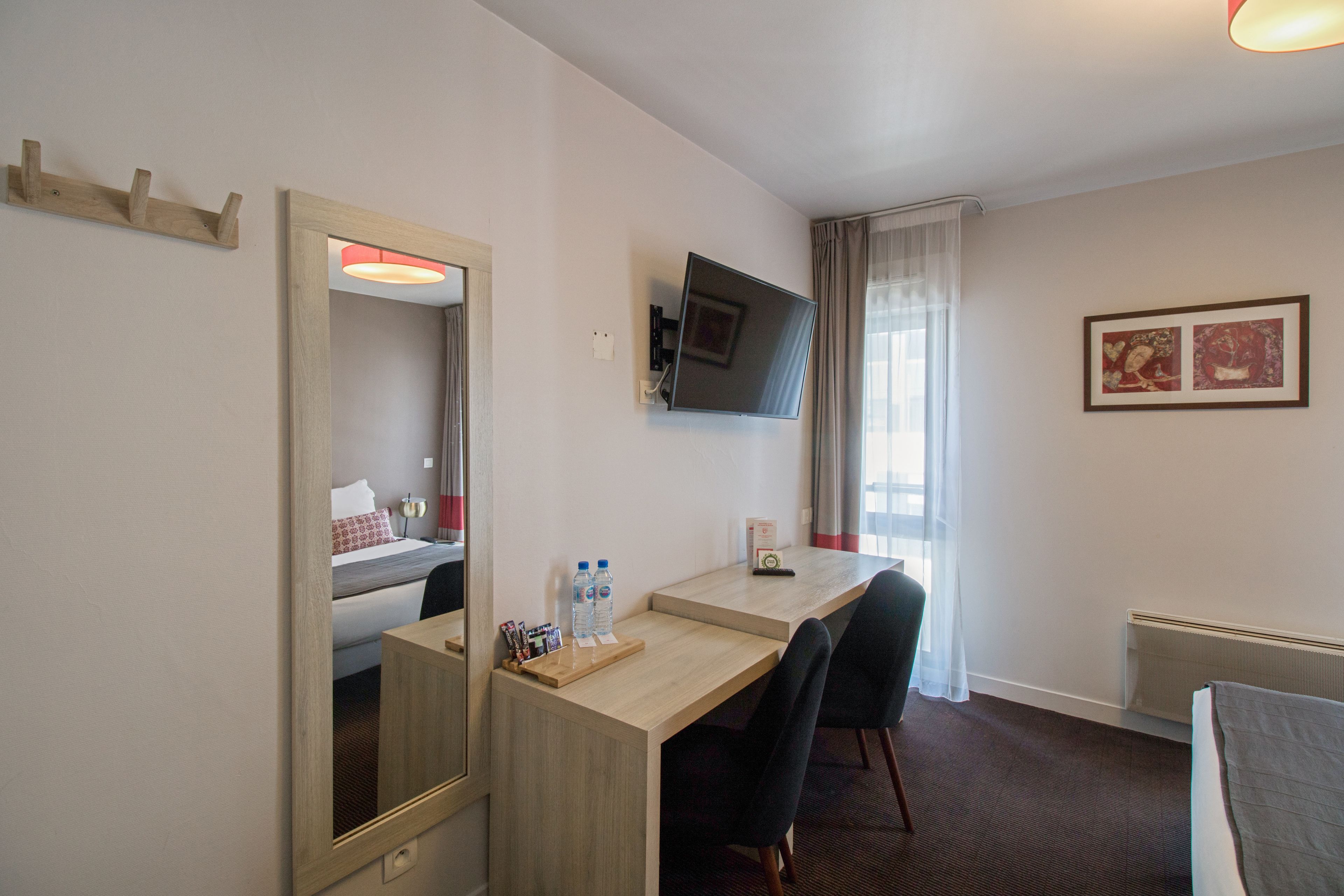 A hotel room with a bed, a desk with two chairs, a TV mounted on the wall, and a window with curtains.