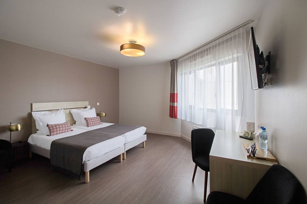 Una habitación de hotel con cama, mesitas de noche, un escritorio, una silla, un televisor montado en la pared y una ventana con cortinas.