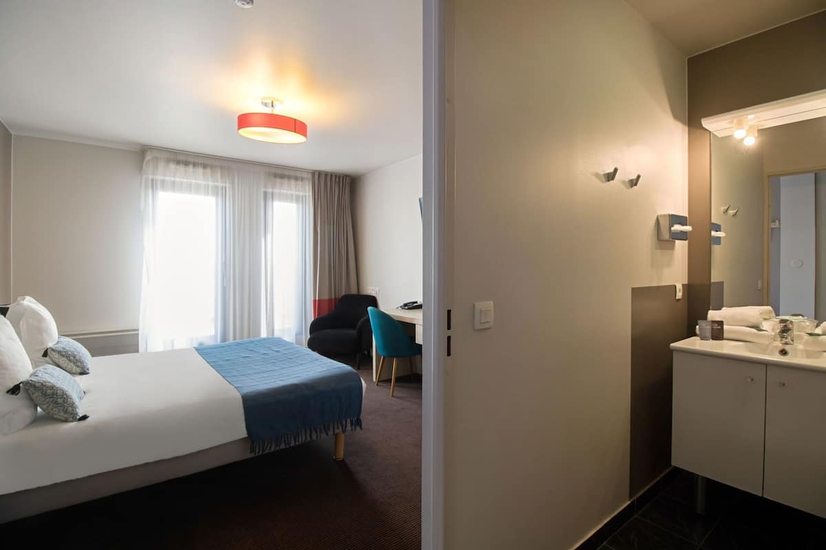 Habitación de hotel con cama, escritorio, silla, baño con espejo y ventana con cortinas.