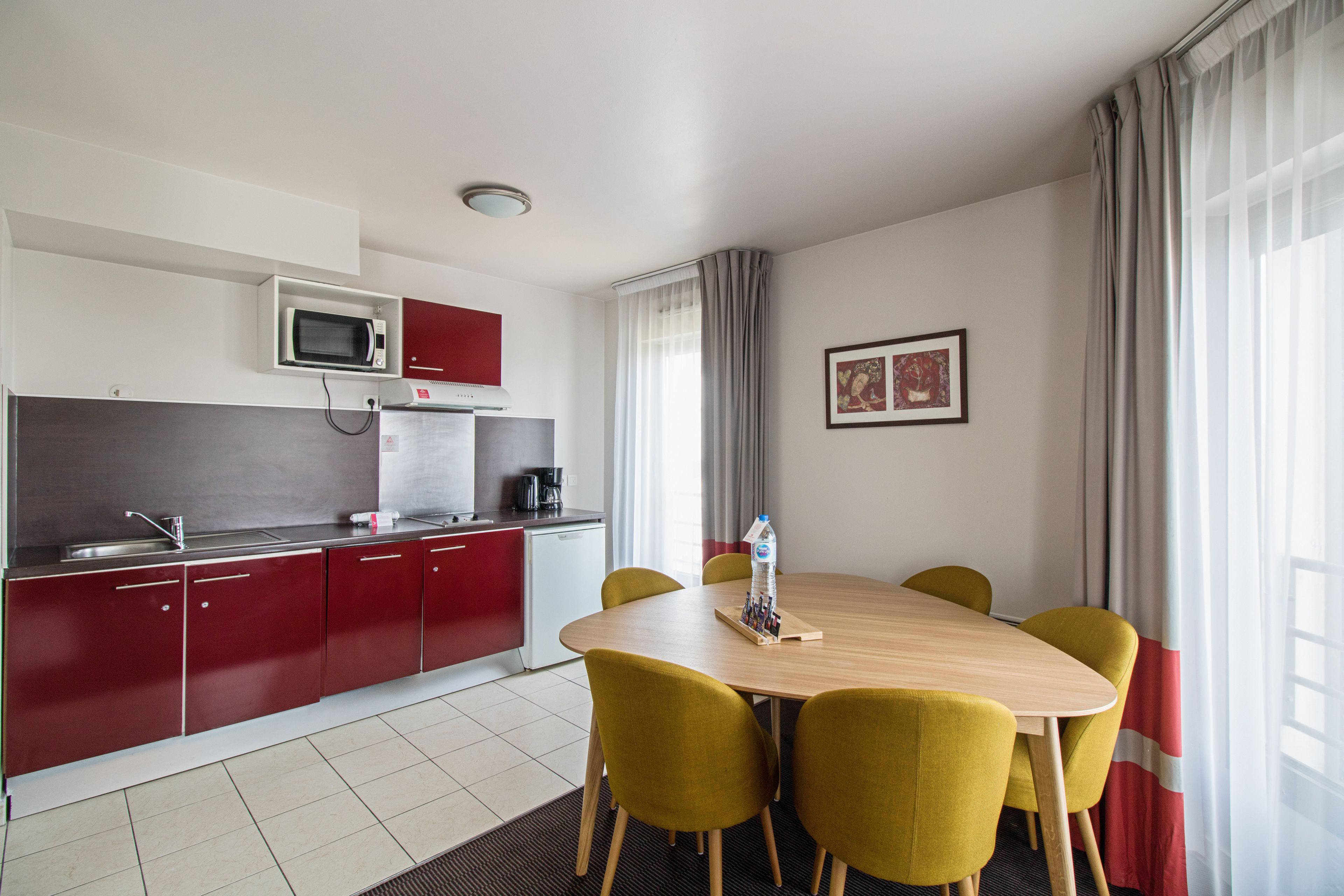 A modern kitchen with red cabinets, a microwave, and a coffee maker. There is a dining table with yellow chairs and a bottle of water.