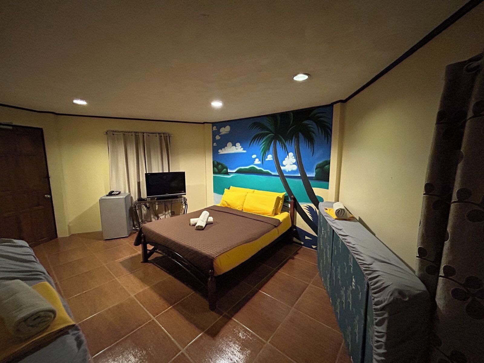 A hotel room with a bed, a television, a desk, and a mural of a beach scene.