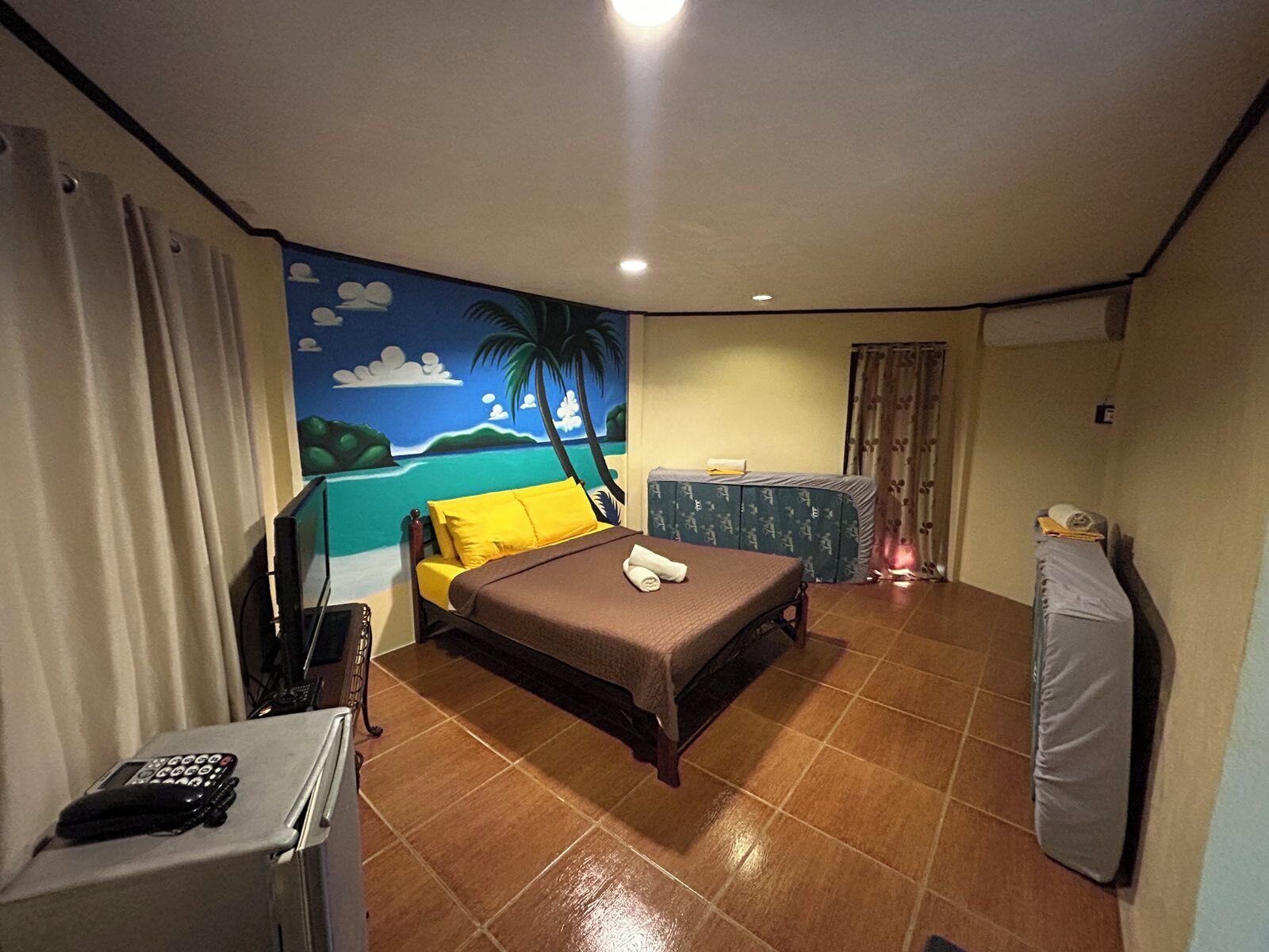 A hotel room with a bed, a desk with a telephone, a small table with a lamp, and a wall mural.