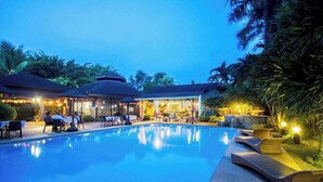 Una piscina con sillas de descanso, un área de estar techada y un restaurante con iluminación exterior.