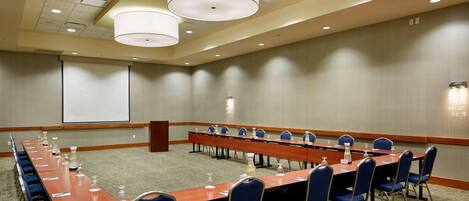Un salón de conferencias con una larga mesa, sillas azules y una pantalla de proyector.