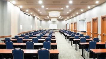 A conference room with rows of tables and chairs, a large screen at the front, and wooden doors along the walls.