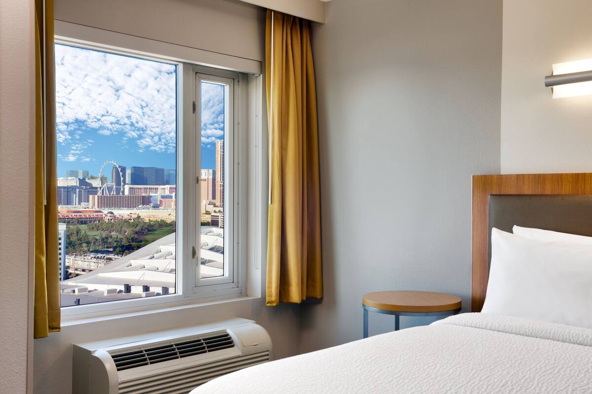 A hotel room with a large window, a bed, a bedside table, and an air conditioning unit.