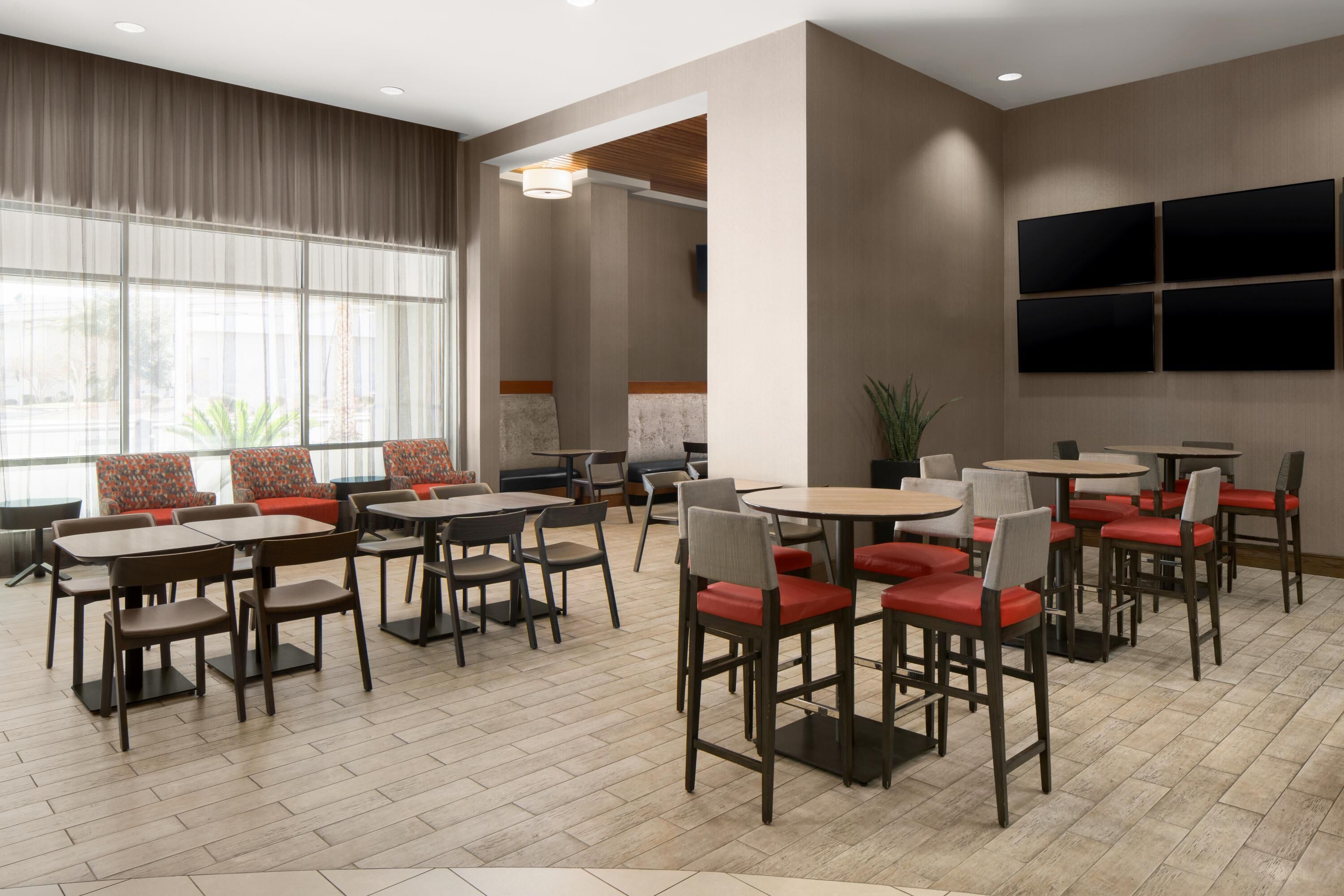 A modern dining area with multiple tables and chairs, large windows, and a flat-screen TV mounted on the wall.