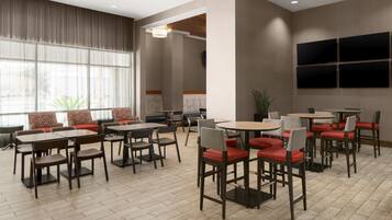 A modern dining area with multiple tables and chairs, large windows, and a flat-screen TV mounted on the wall.