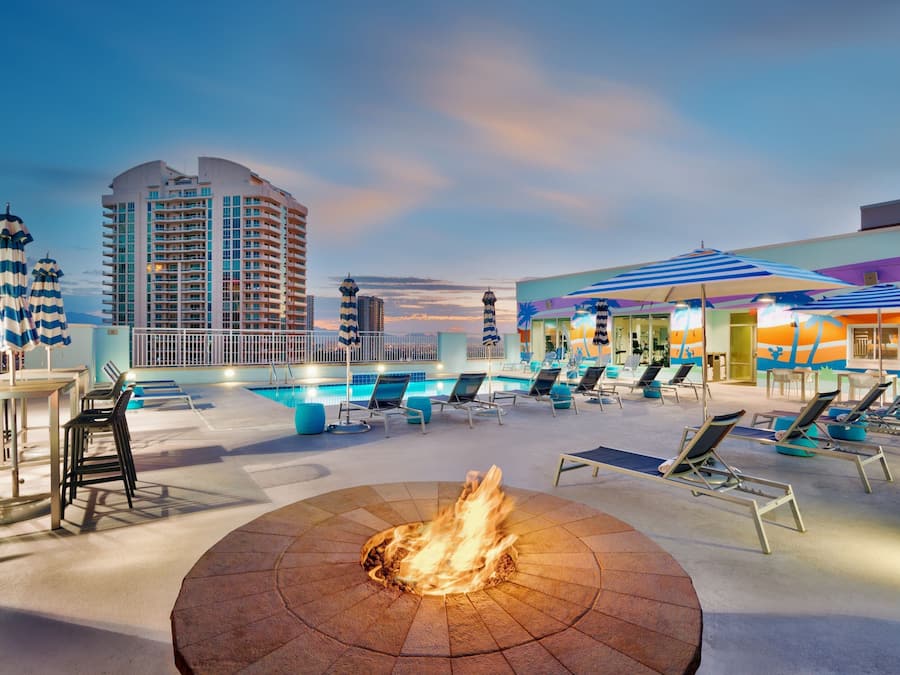 A rooftop pool area with lounge chairs, a fire pit, and a view of a tall hotel building.