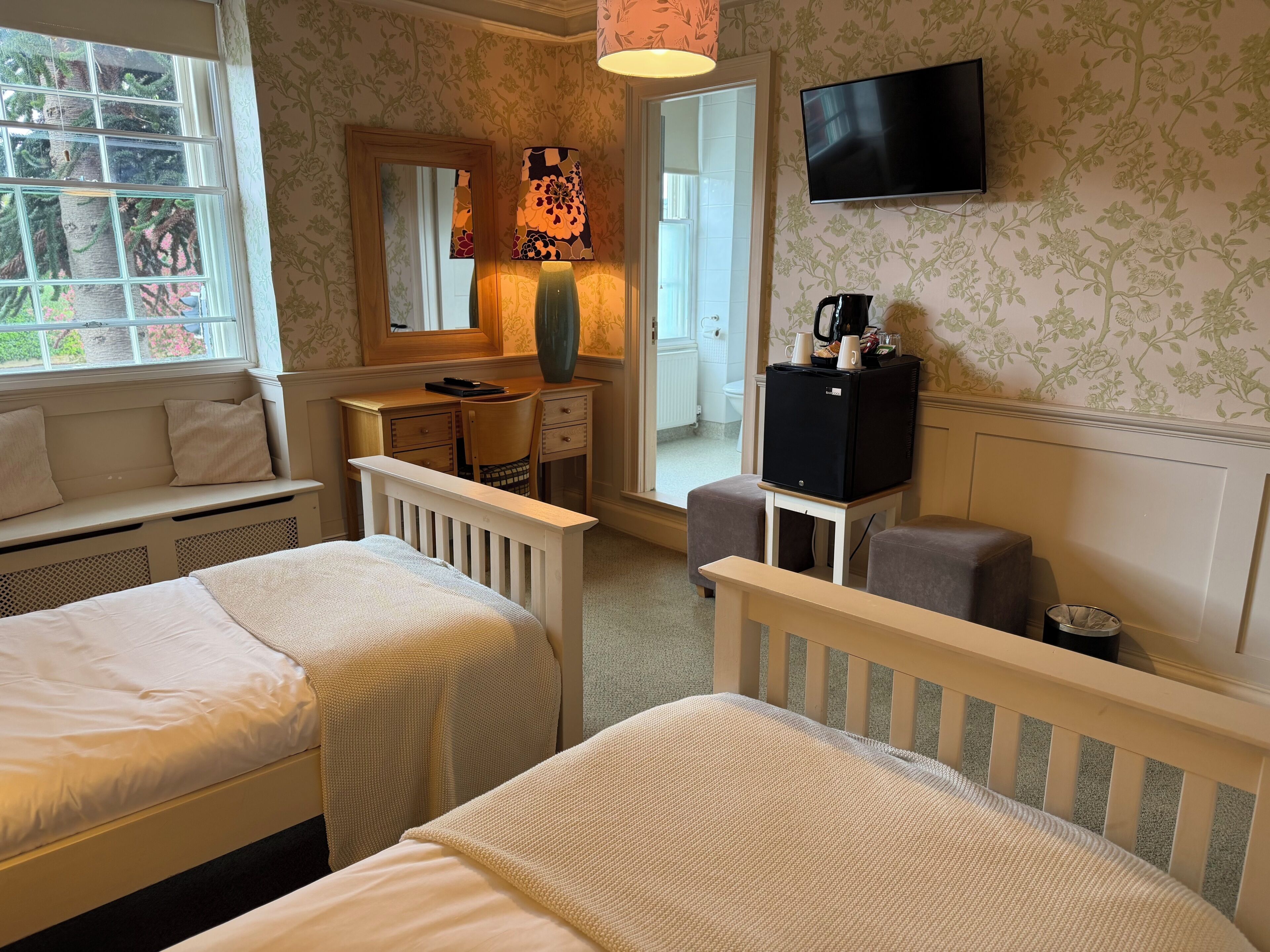 A hotel room with two beds, a desk, a chair, a television, and a window with a view of greenery.