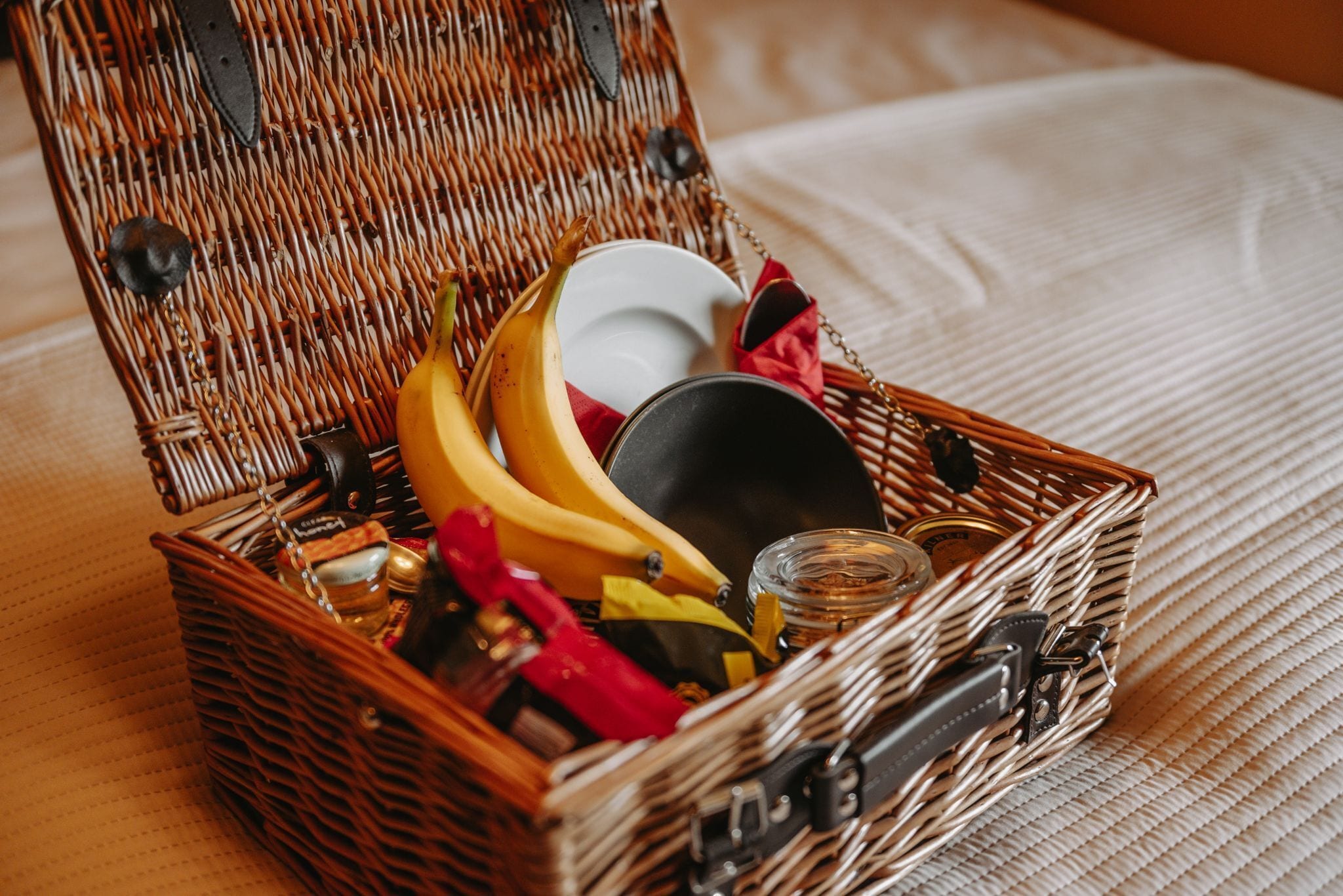 A wicker basket with a metal clasp, containing bananas, a white plate, a red napkin, and a small jar.