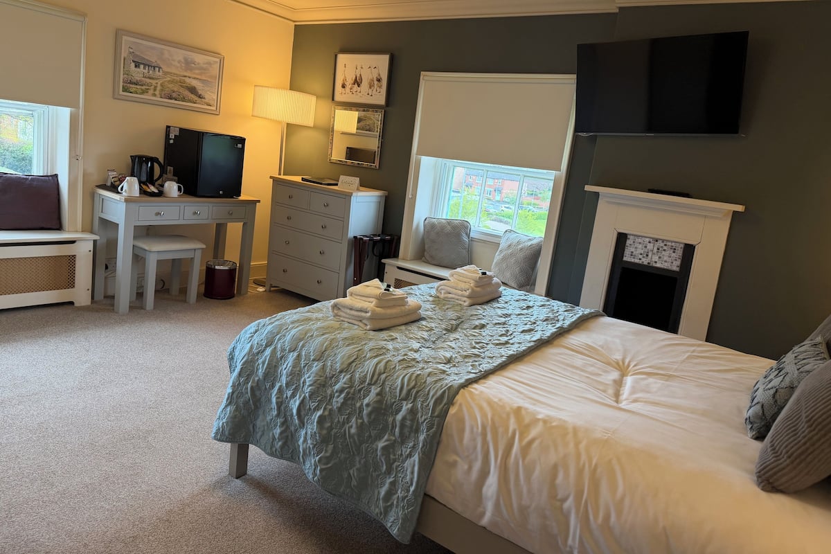 A bedroom with a bed, a desk, a television, a fireplace, and a window.