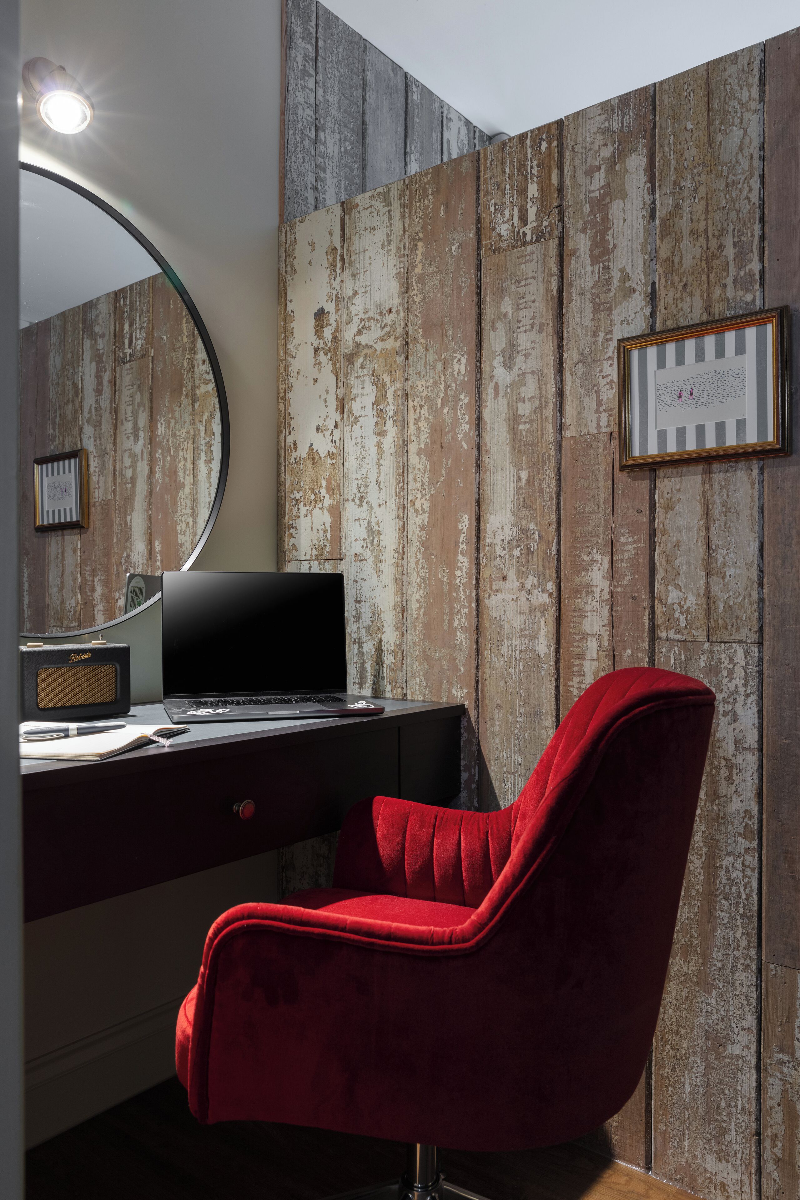 Habitación con paneles de madera, un escritorio con una computadora portátil, una silla roja, un espejo redondo y un cuadro enmarcado.