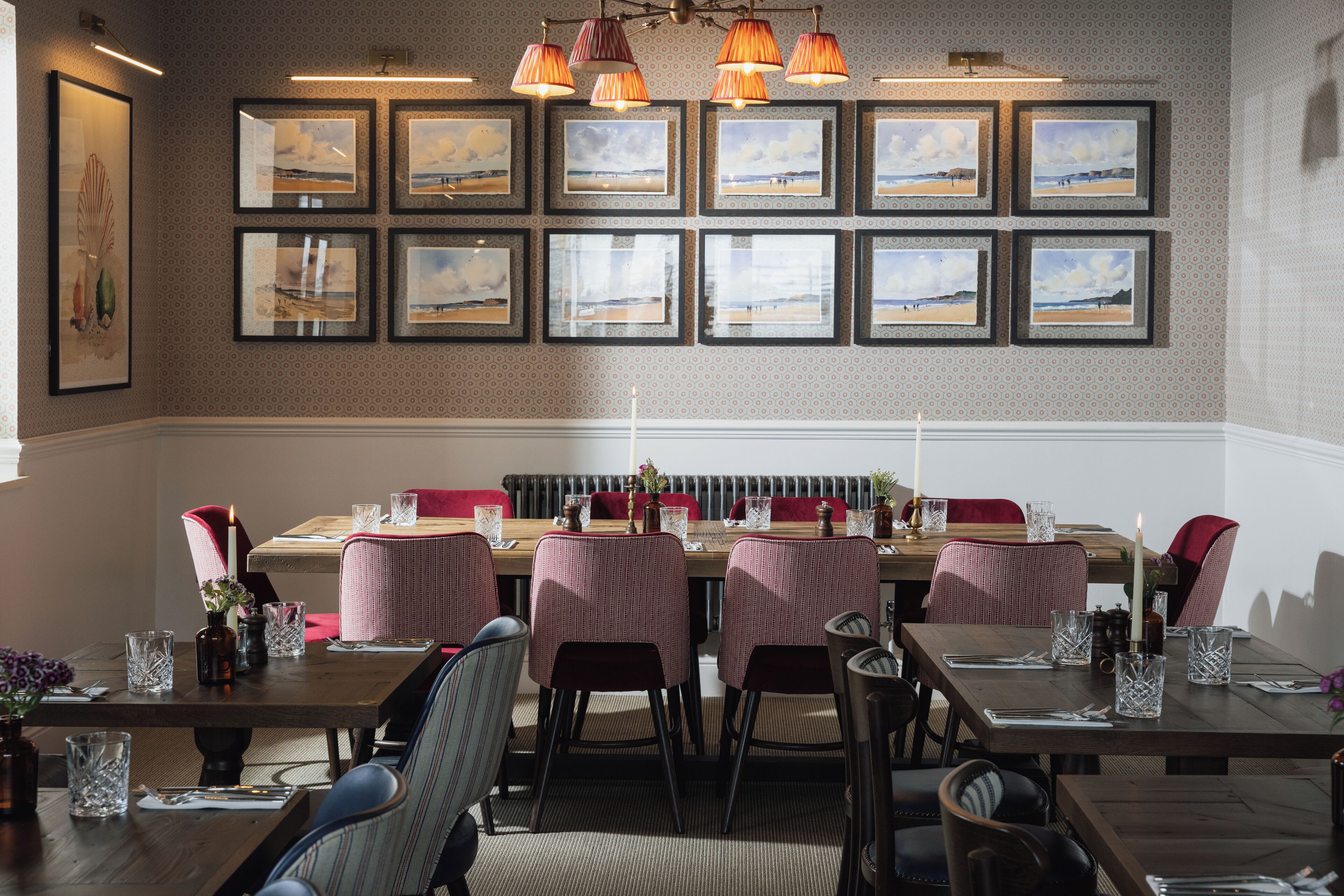 A dining area with a long table set for multiple guests, framed coastal landscape pictures on the wall, and a patterned wallpaper design.