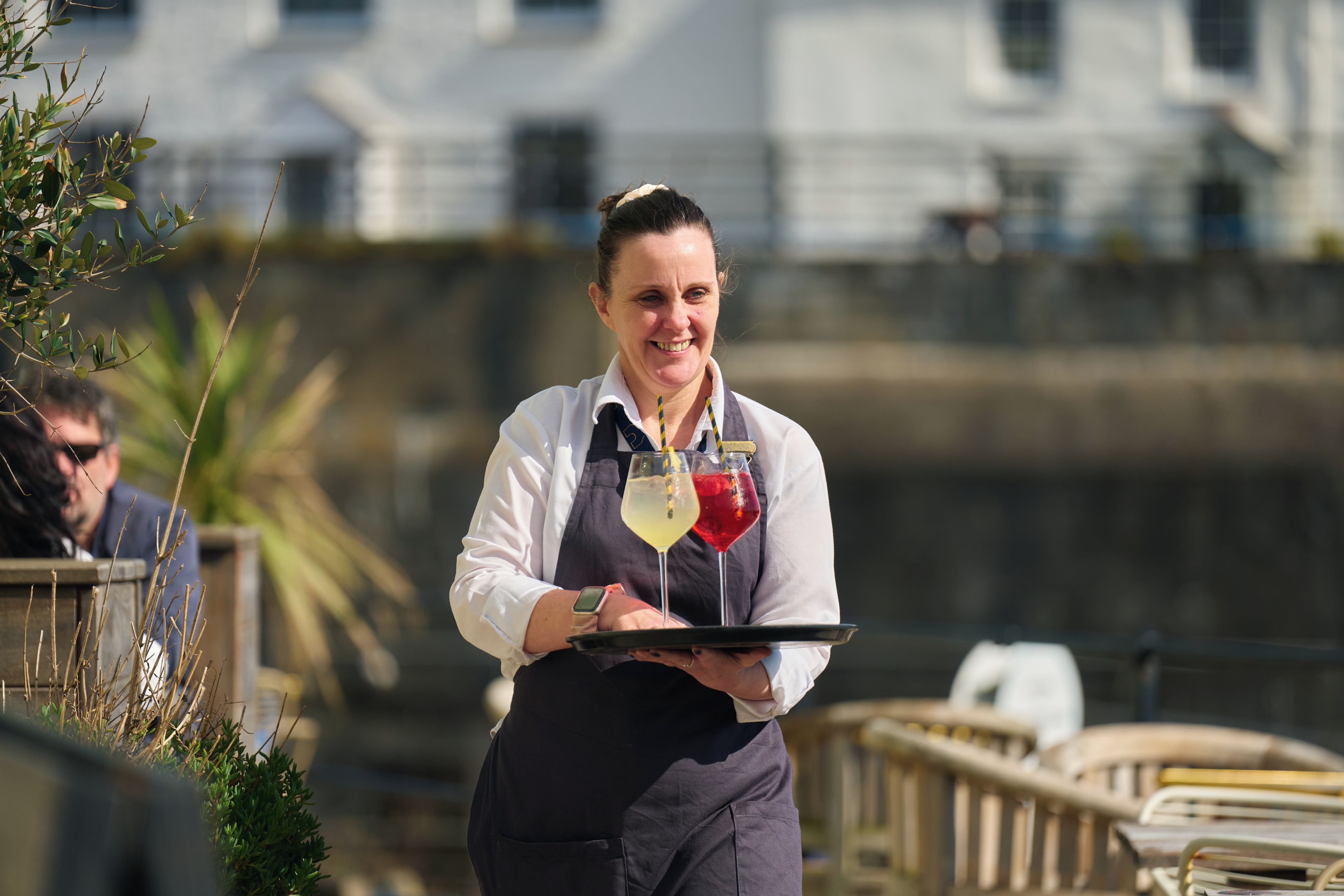 A person carrying a tray with drinks, standing outdoors with a railing and greenery in the background.