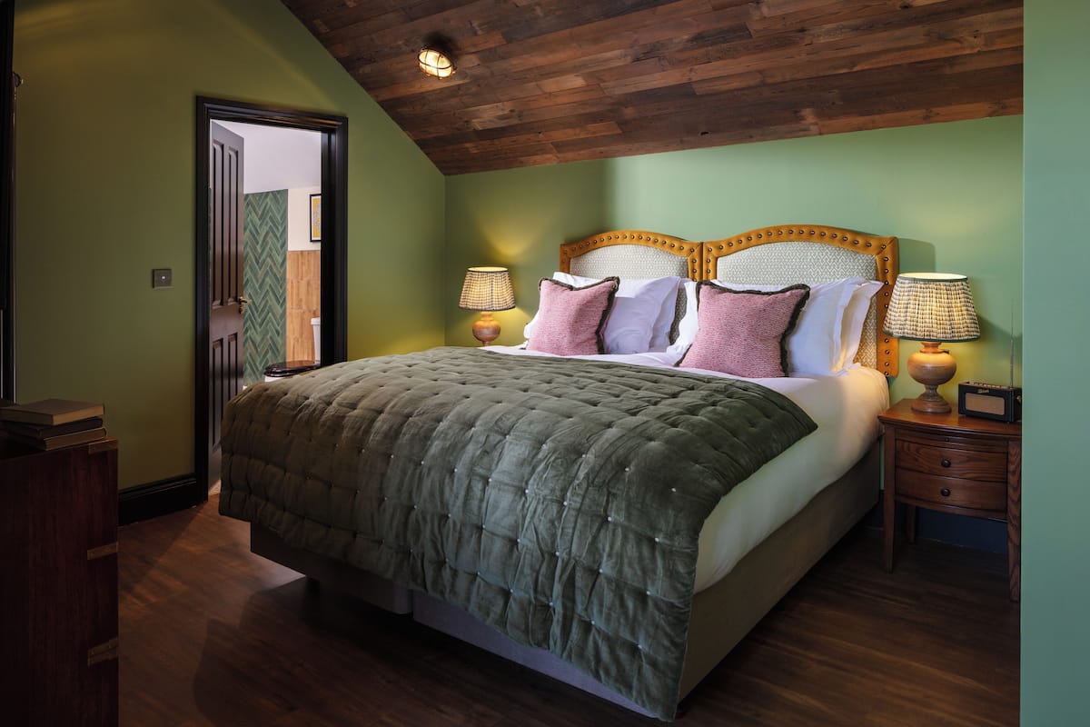 Un dormitorio con una cama grande, cabecera de madera, dos mesitas de noche con lámparas y una puerta que da a otra habitación.