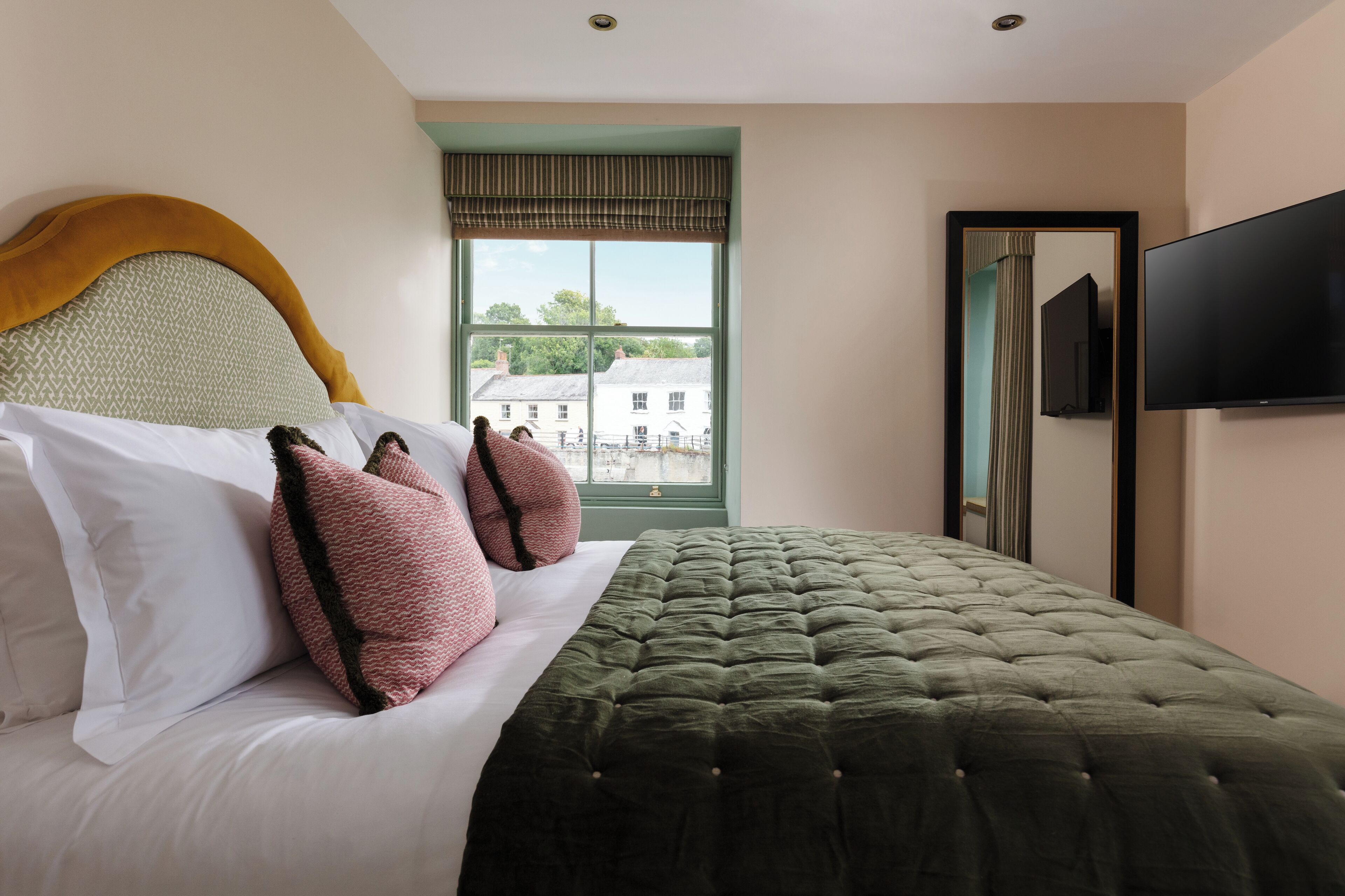 A neatly made bed with a green quilt, white pillows, and two pink pillows. A large window with a view of buildings is visible.