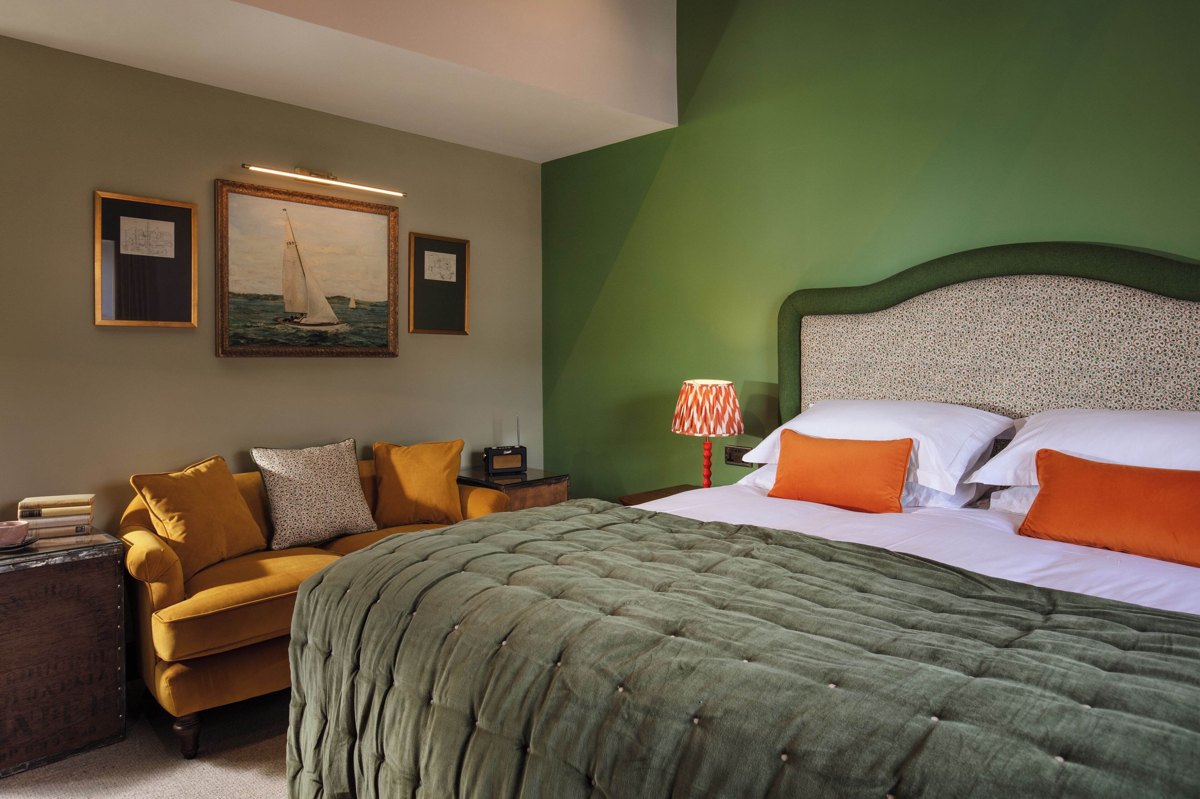 Une chambre d’hôtel avec un mur vert, un lit avec des coussins blancs et orange, une couvre-lit matelassée verte, un fauteuil jaune, une petite table avec une radio et deux tableaux encadrés accrochés au mur.